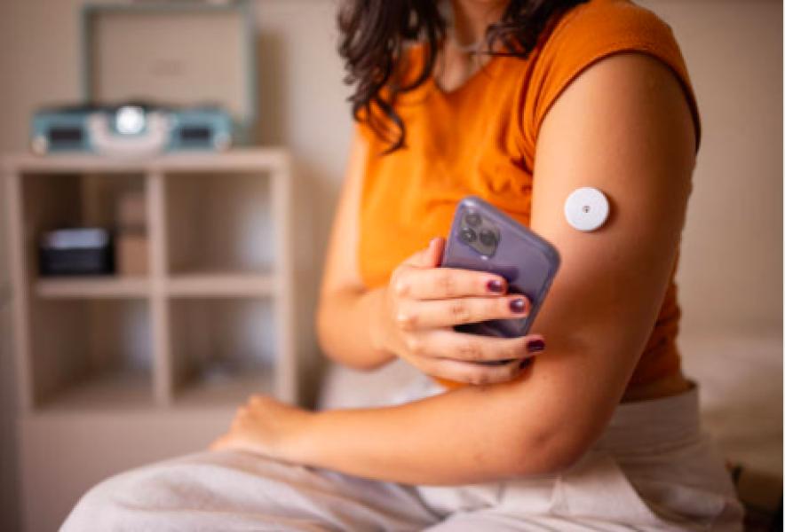 Woman wearing orange shirt displaying continuous glucose monitoring device on arm while holding blood glucose meter