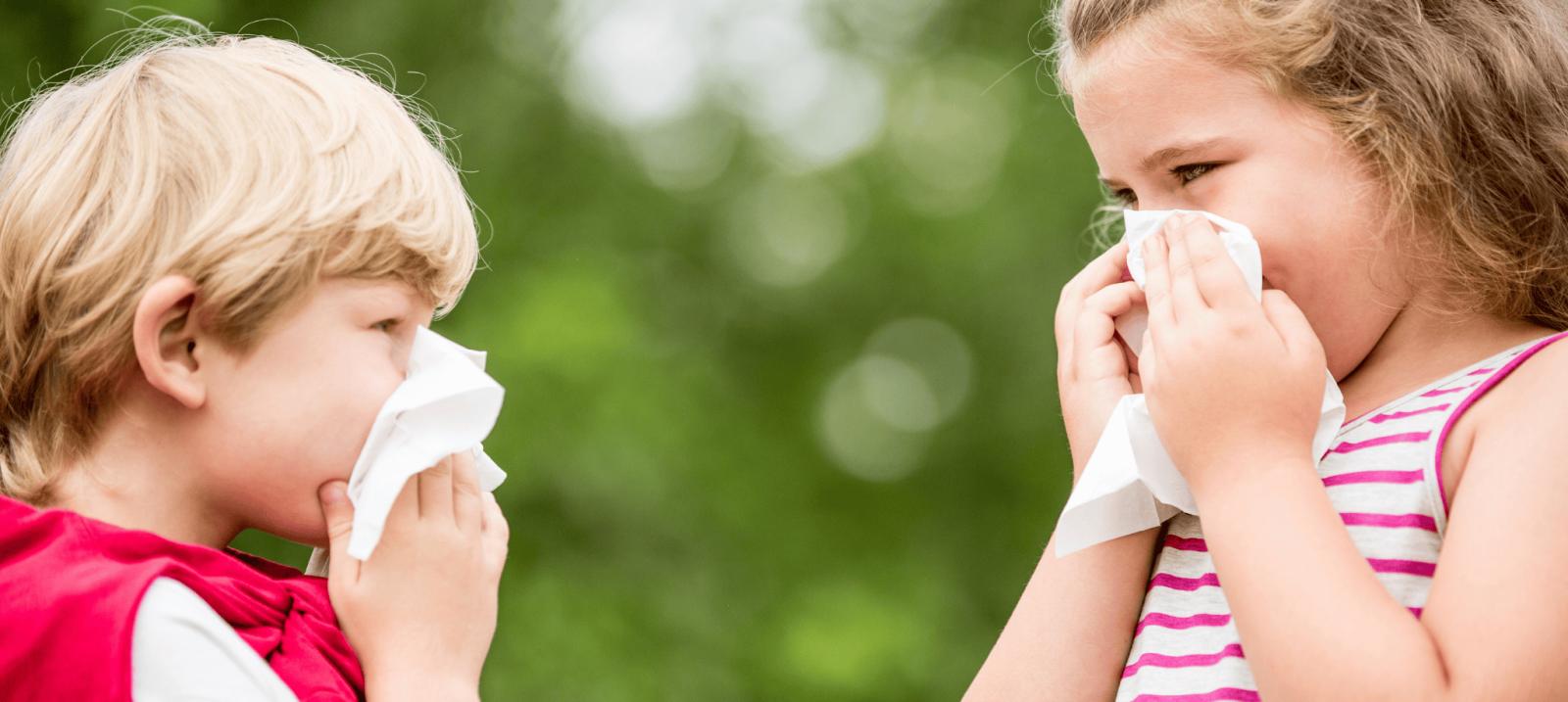 Two young children outdoors holding tissues to their noses, showing signs of allergies or cold symptoms
